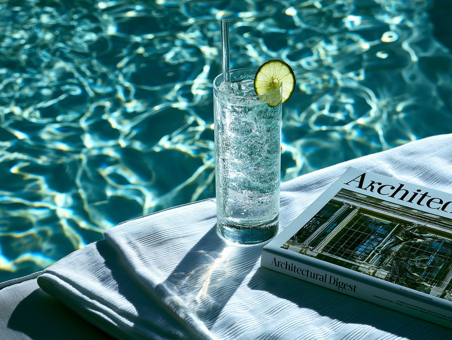 A close-up of a tall glass filled with a clear icy drink garnished with a lime slice and a straw, placed on a folded white towel next to an Architectural Digest magazine, sitting by a rippling blue pool.