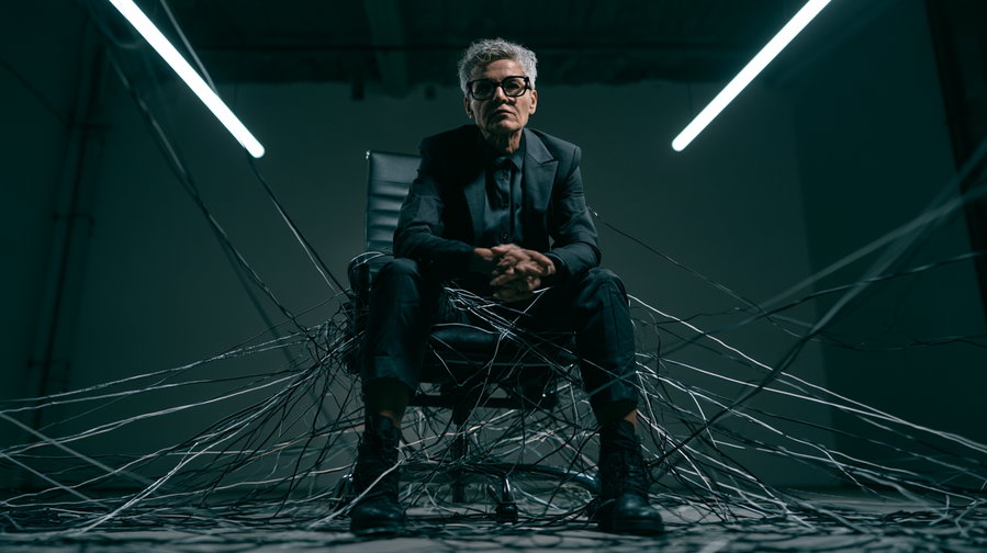 A person with short gray hair and glasses wears a dark suit and boots, sitting on a chair surrounded and entangled by many wires in a dimly lit room with overhead lights.