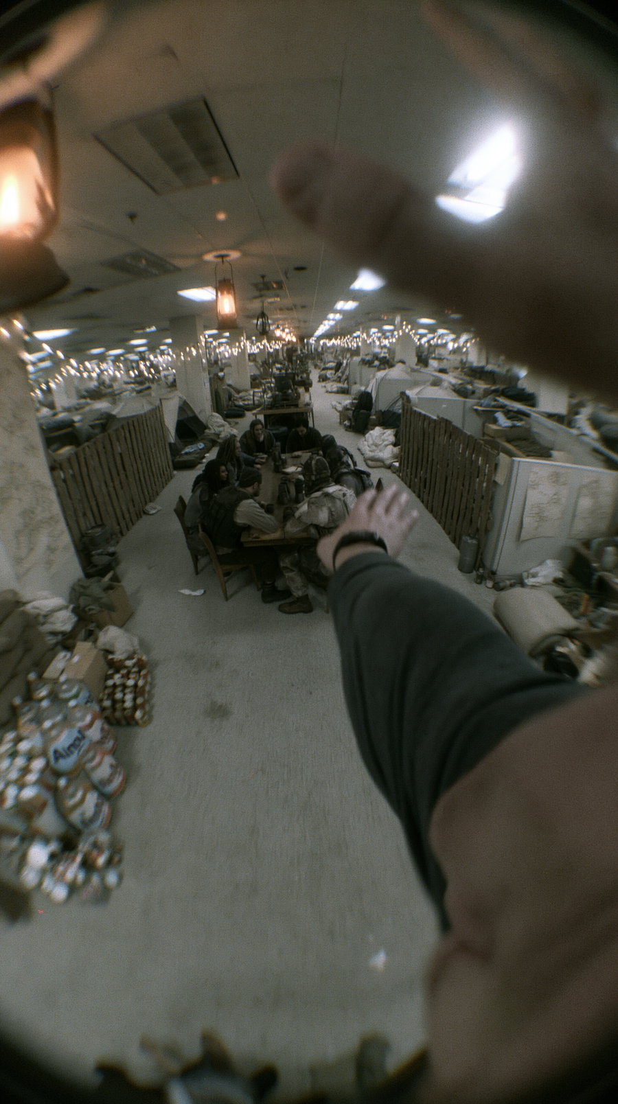 A group of people sitting at a table in a large indoor space filled with beds and supplies, viewed through a circular fisheye lens with a hand partially blocking the view
