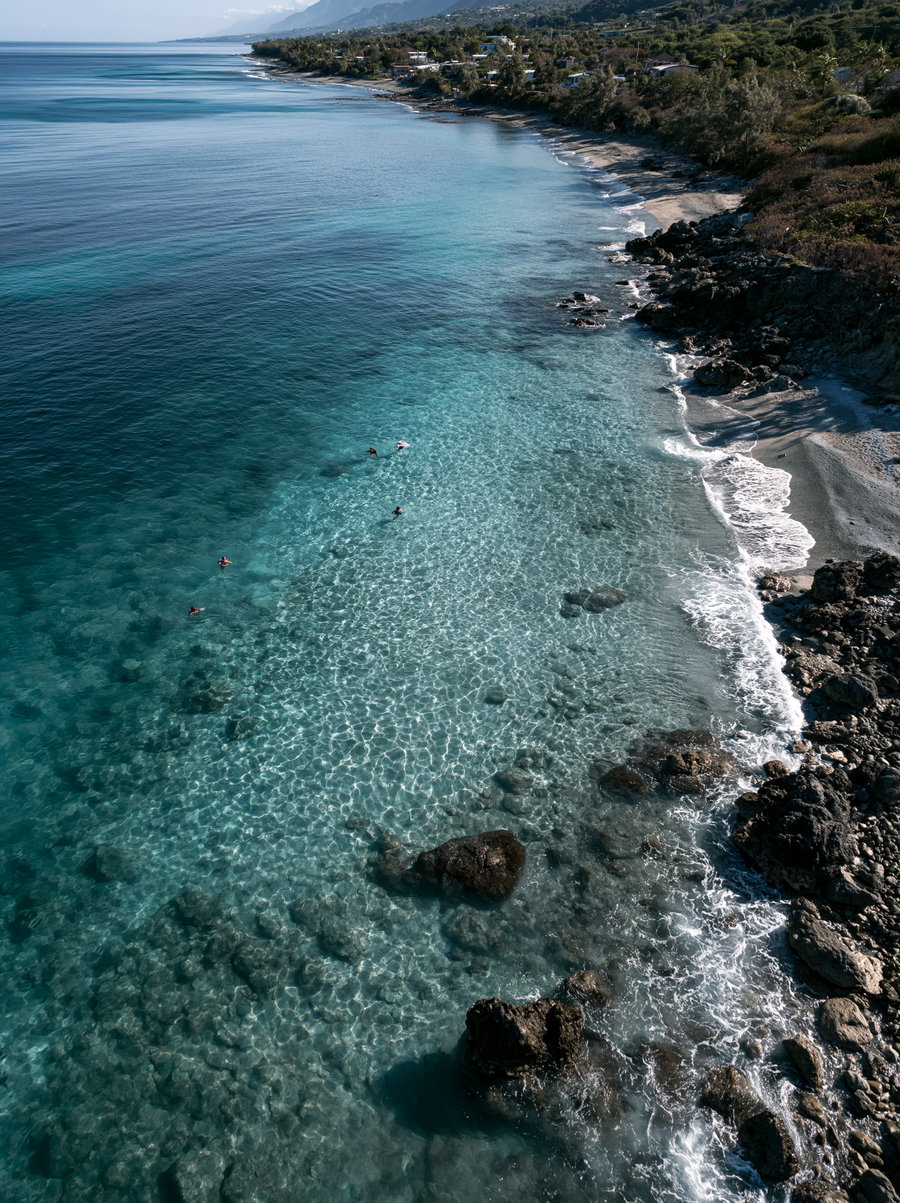 A clear blue ocean with visible rocks beneath the water, a rocky shoreline with small waves, and several people swimming near the coast under a distant mountain and green trees.