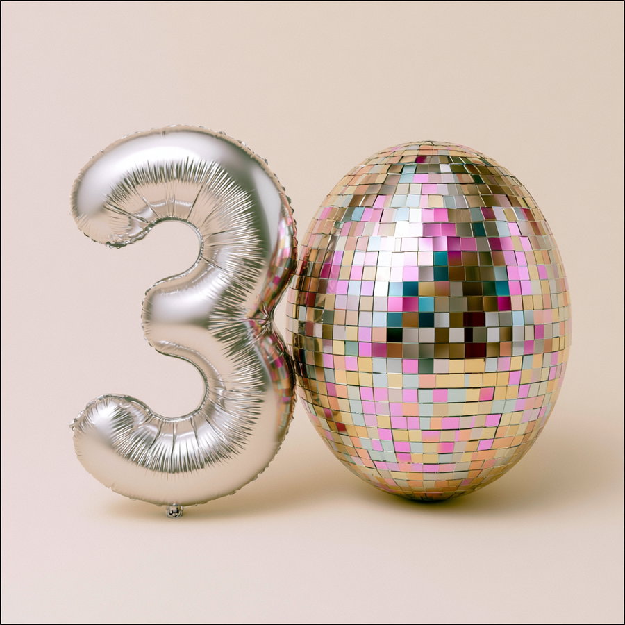 Silver number three balloon next to a shiny egg covered in small mirrored tiles reflecting pink, gold, and green colors on a beige background