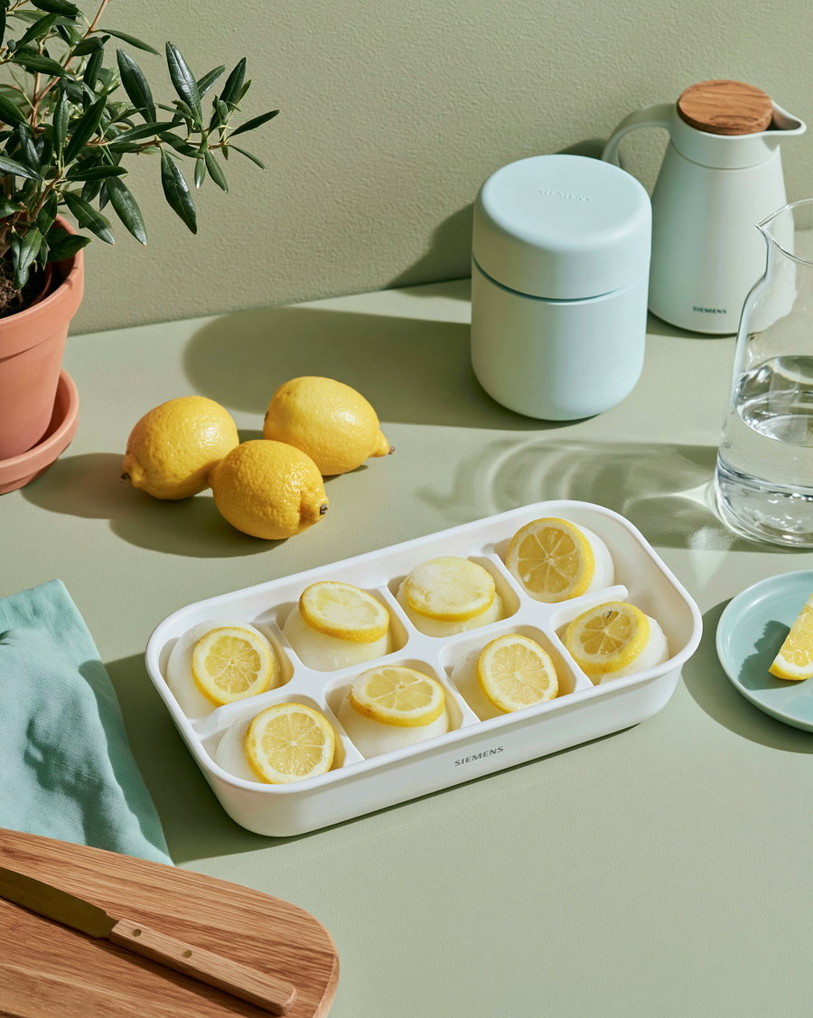 A white ice tray with lemon slices frozen in ice cubes placed on a pale green countertop, surrounded by a potted plant, three whole lemons, a white pitcher, a glass pitcher with water, a wooden cutting board with a knife, and a small green plate with a lemon wedge