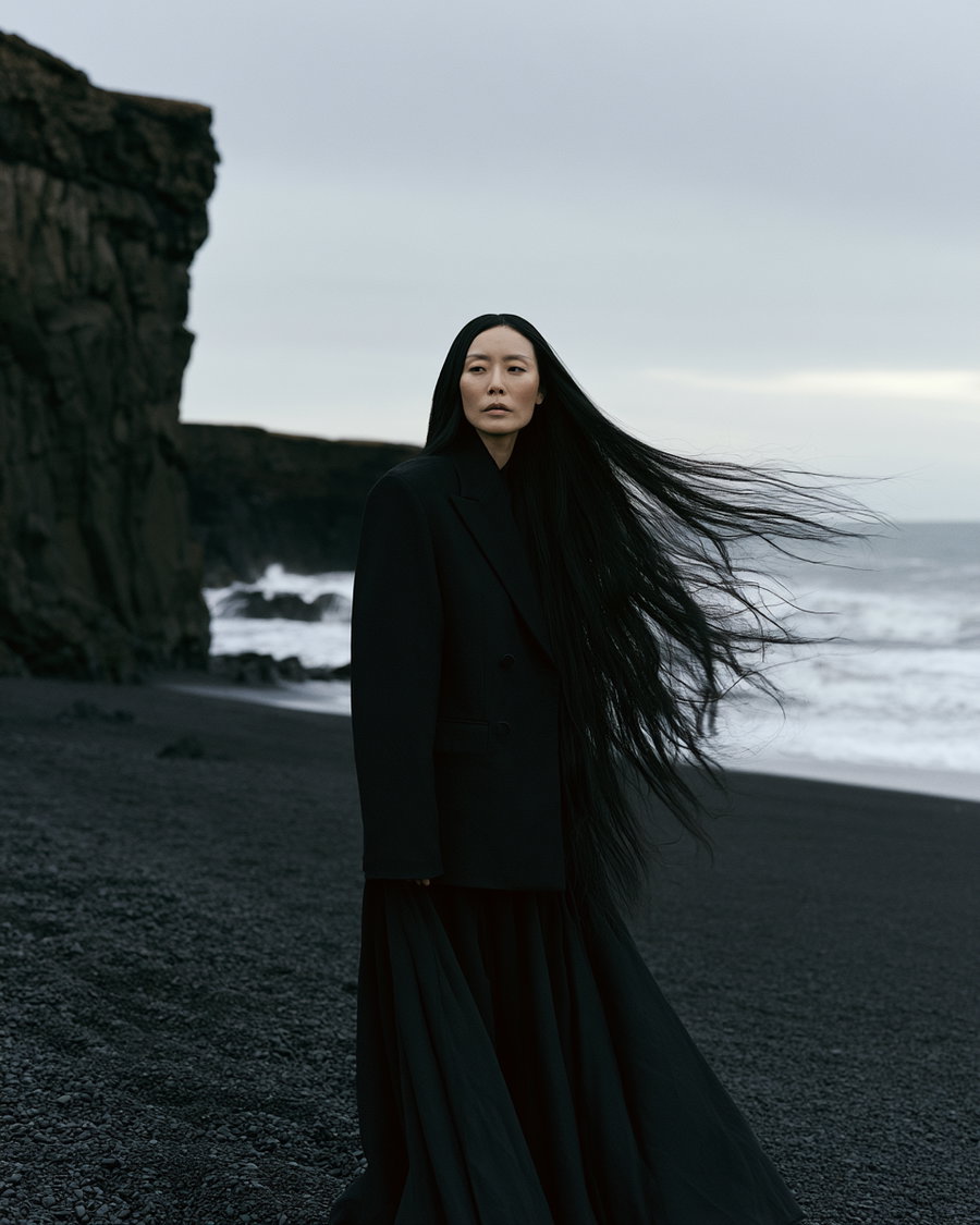 A woman with very long black hair standing on a black rock beach with cliffs in the background and ocean waves nearby, wearing a long black coat and skirt
