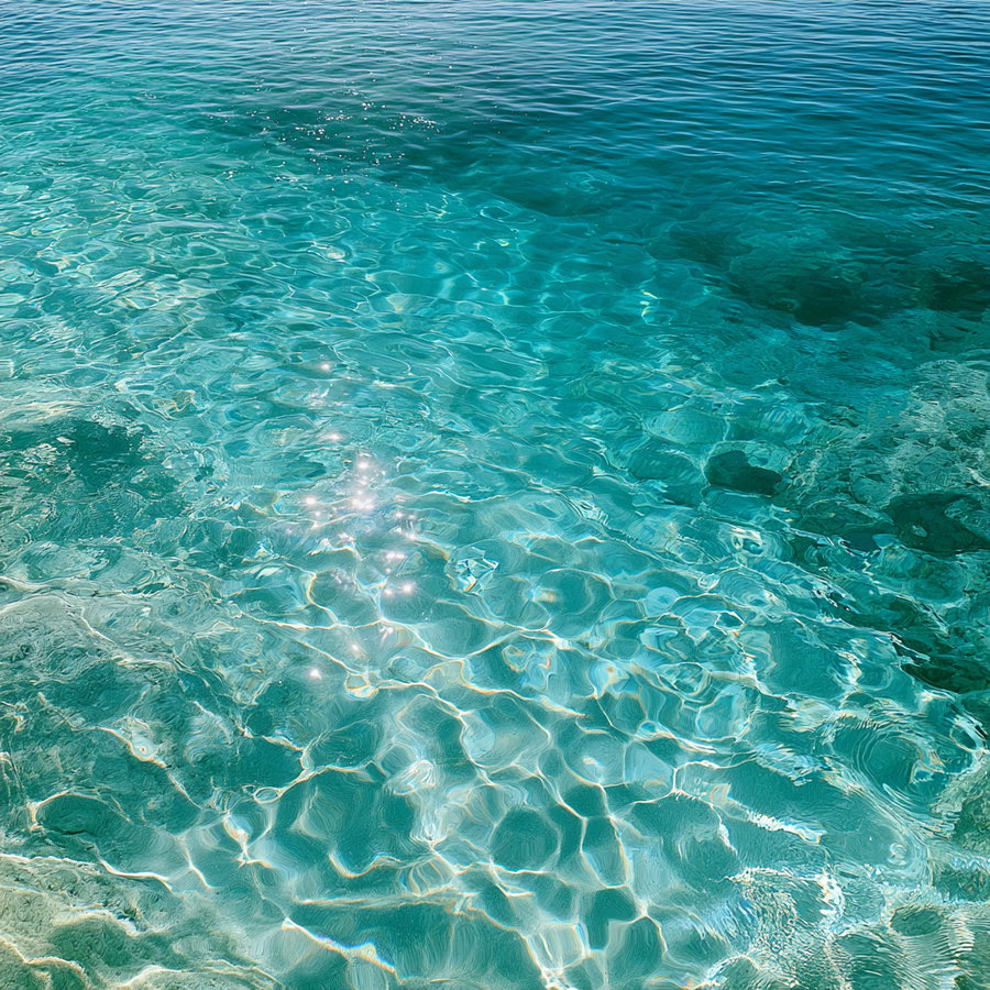 Close-up view of crystal clear turquoise ocean water with sunlight reflecting and sparkling on the surface, showing underwater rocks and gentle waves