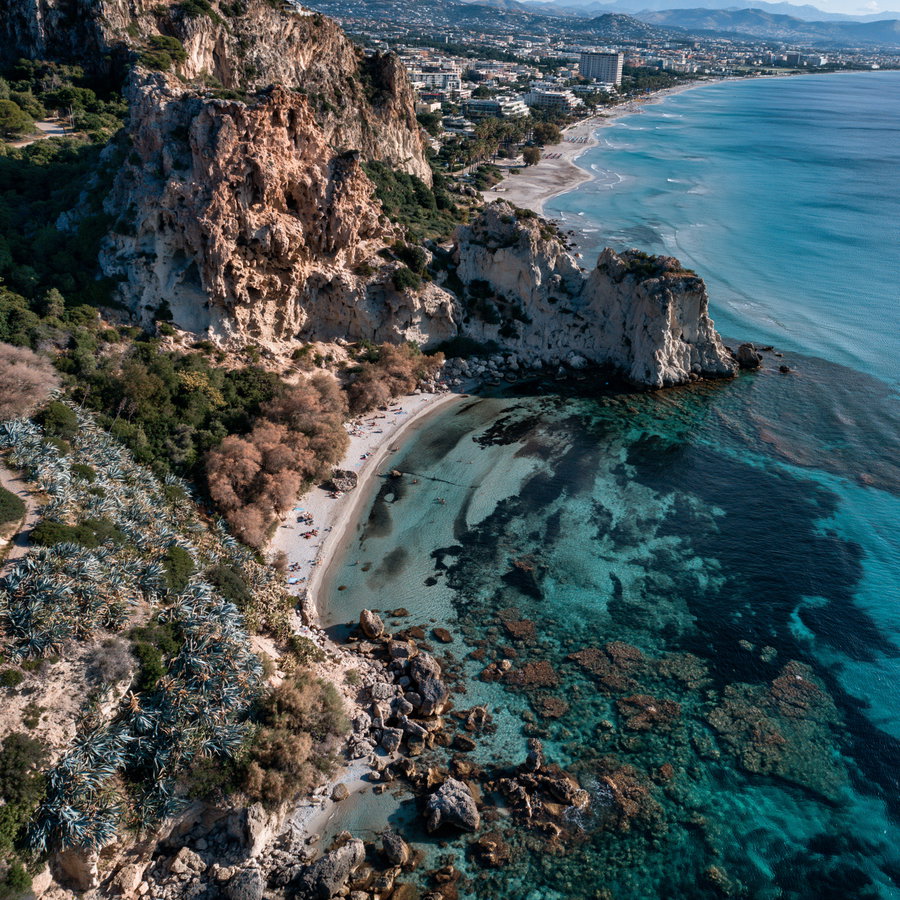 Aerial view of a coastal area featuring rocky cliffs, sandy beach with some people, dense vegetation, and clear blue ocean water with visible underwater rocks.