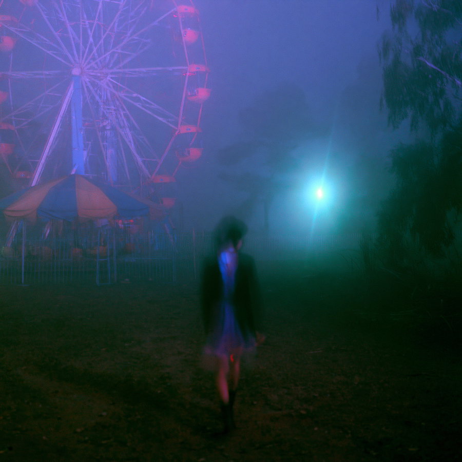 A blurred figure walking in a foggy area with a lit ferris wheel and a bright light in the background, surrounded by dark trees.