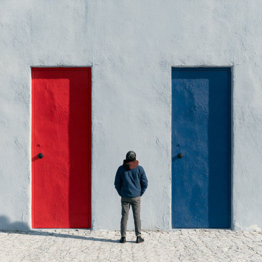 A man in casual attire stands facing two doors, one red and one blue, set against a textured grey wall.