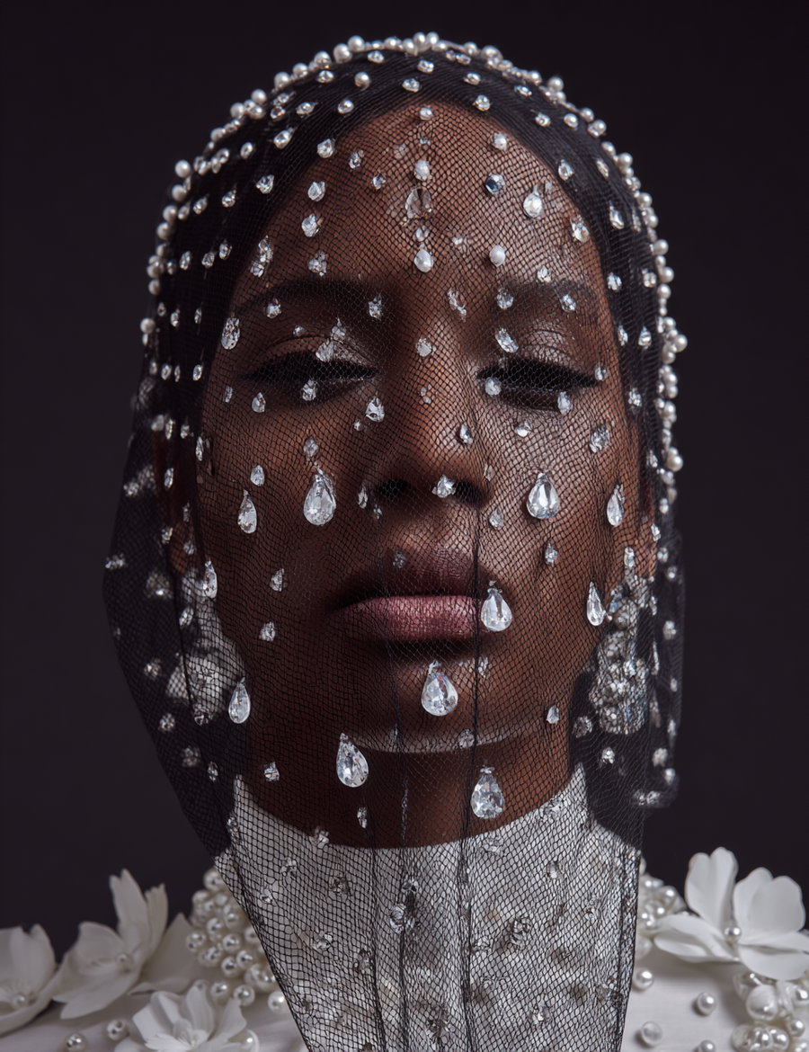 Close-up portrait of a woman wearing a black net veil decorated with teardrop-shaped crystals, a white high-collar garment with pearl and crystal embellishments, and white flowers around her neck on a dark background