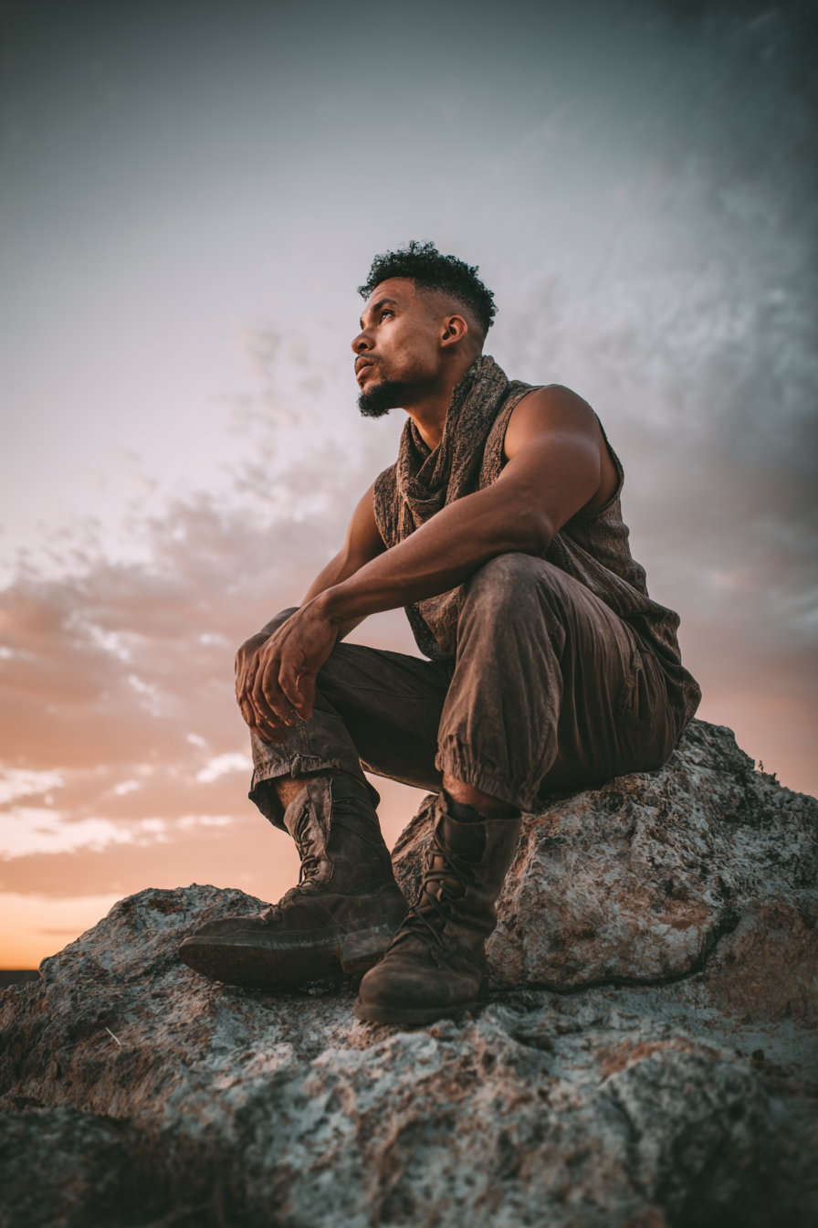 A man with curly hair and a beard wearing a sleeveless top, scarf, cargo pants, and boots sits on a rock looking thoughtfully into the distance during sunset.