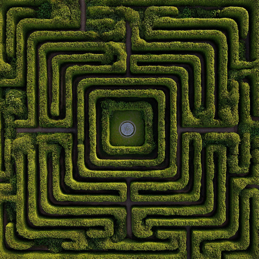 Top-down view of a large, intricate green hedge maze with pathways leading toward a central square area containing a round fountain