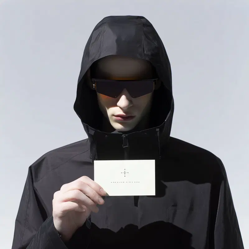 Minimalist portrait of a man in black hood and large sunglasses holding a card