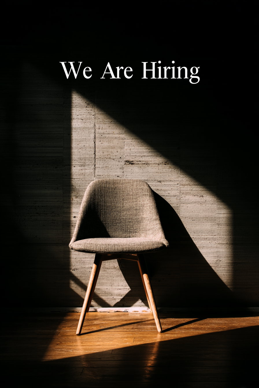 A single wooden-legged chair with fabric seat and backrest stands on a wooden floor, illuminated by a triangular shaft of sunlight against a textured wall, with the text We Are Hiring above it.