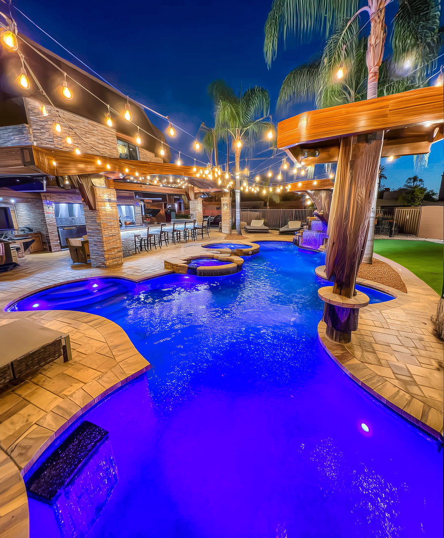Nighttime view of a backyard pool with blue underwater lights, illuminated bar with high chairs, string lights overhead, palm trees, and lounge chairs on the side.